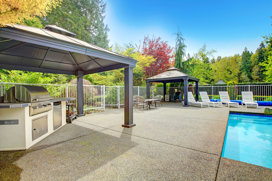 Spacious backyard with a swimming pool, lounge chairs, two covered patios, outdoor grill, and patio furniture set on a concrete pool deck, surrounded by trees and greenery under a clear blue sky.