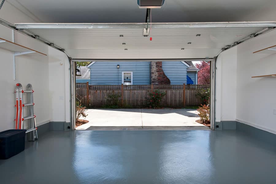 View from inside a clean, empty garage with a shiny gray garage floor concrete. The garage door is open, showing a driveway, wooden fence, and blue house with chimney outside—a great example of concrete options for Michigan homeowners. Shelves and tools line the walls.