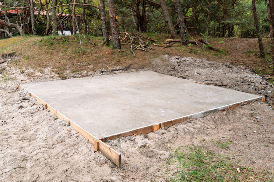 A freshly poured concrete pad forms a flat rectangular foundation for a future backyard shed, surrounded by sand, grass, and trees in an outdoor, forested area. Wooden boards frame the edges of the outbuilding site.