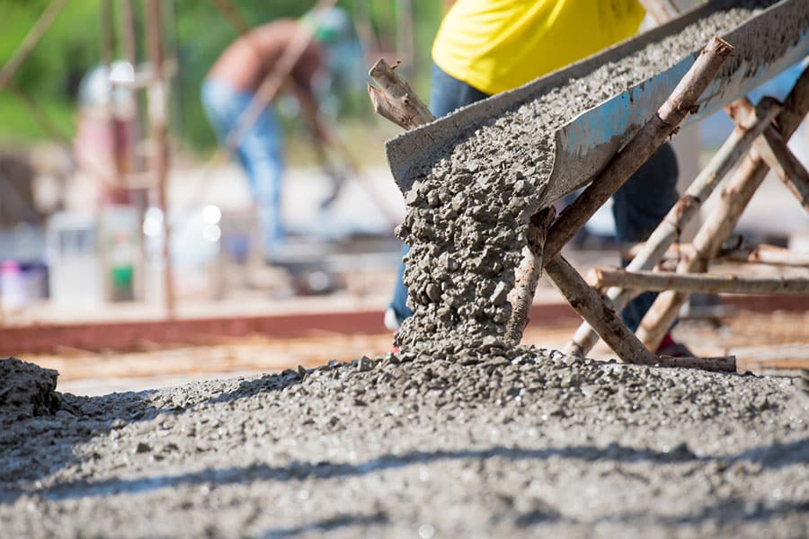 Wet concrete pours from a chute onto a surface at a construction site during peak concrete season, with workers and scaffolding visible in the blurred background.