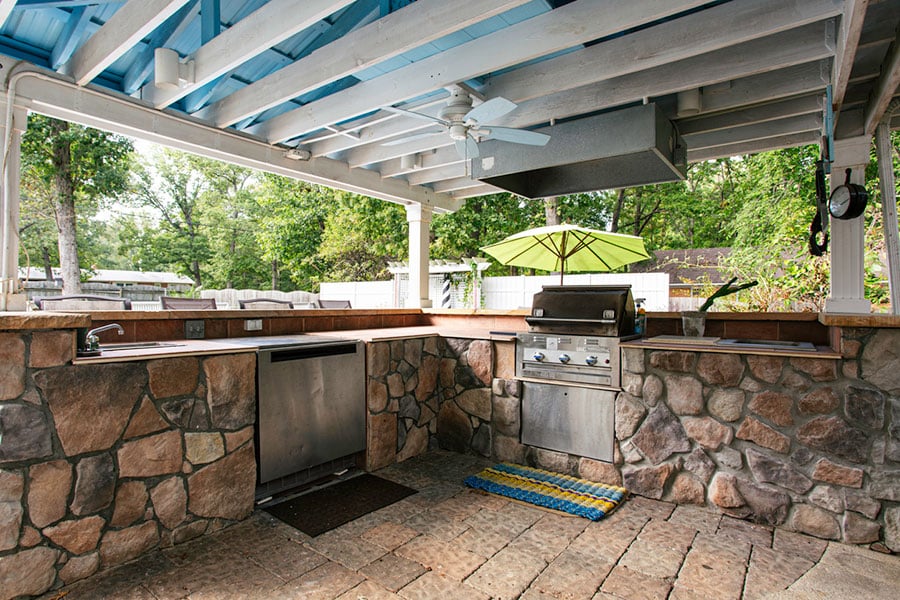 Outdoor kitchen with stone counters, stainless steel grill, and sink under a covered patio with a ceiling fan. A cozy firepit adds warmth nearby, perfect for gatherings. Green umbrella and trees are visible in the background. Multicolored mat is on the floor.