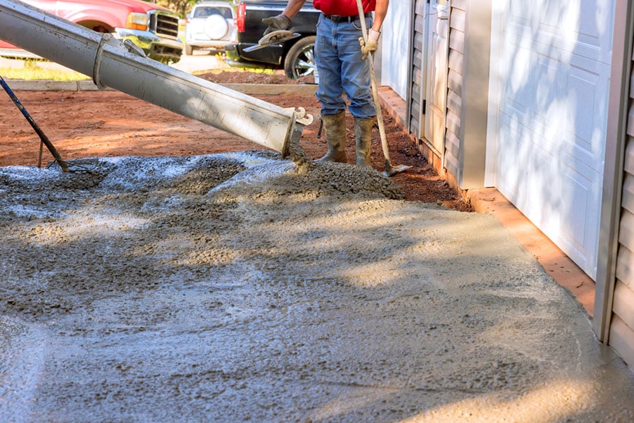 A worker in boots and gloves uses a tool to spread wet concrete pouring from a chute during a driveway installation.